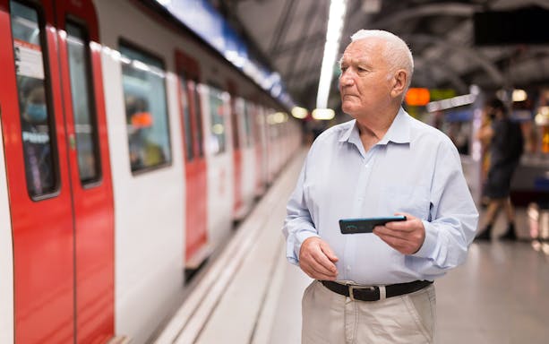 Elderly man holding a phone at a train station platform.