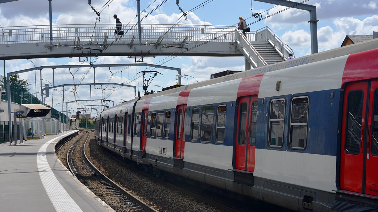 Paris RER train at a station with passengers on an overhead footbridge.