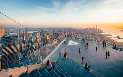 Observation deck view of New York City skyline at sunset, featuring visitors and glass railings.