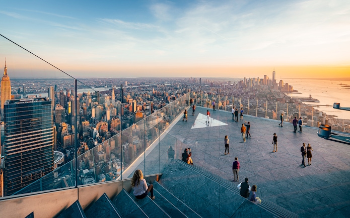 Observation deck view of New York City skyline at sunset, featuring visitors and glass railings.