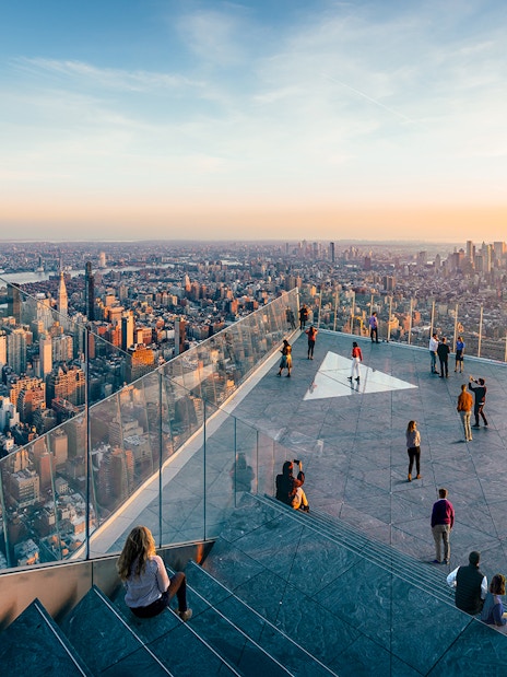 Observation deck view of New York City skyline at sunset, featuring visitors and glass railings.