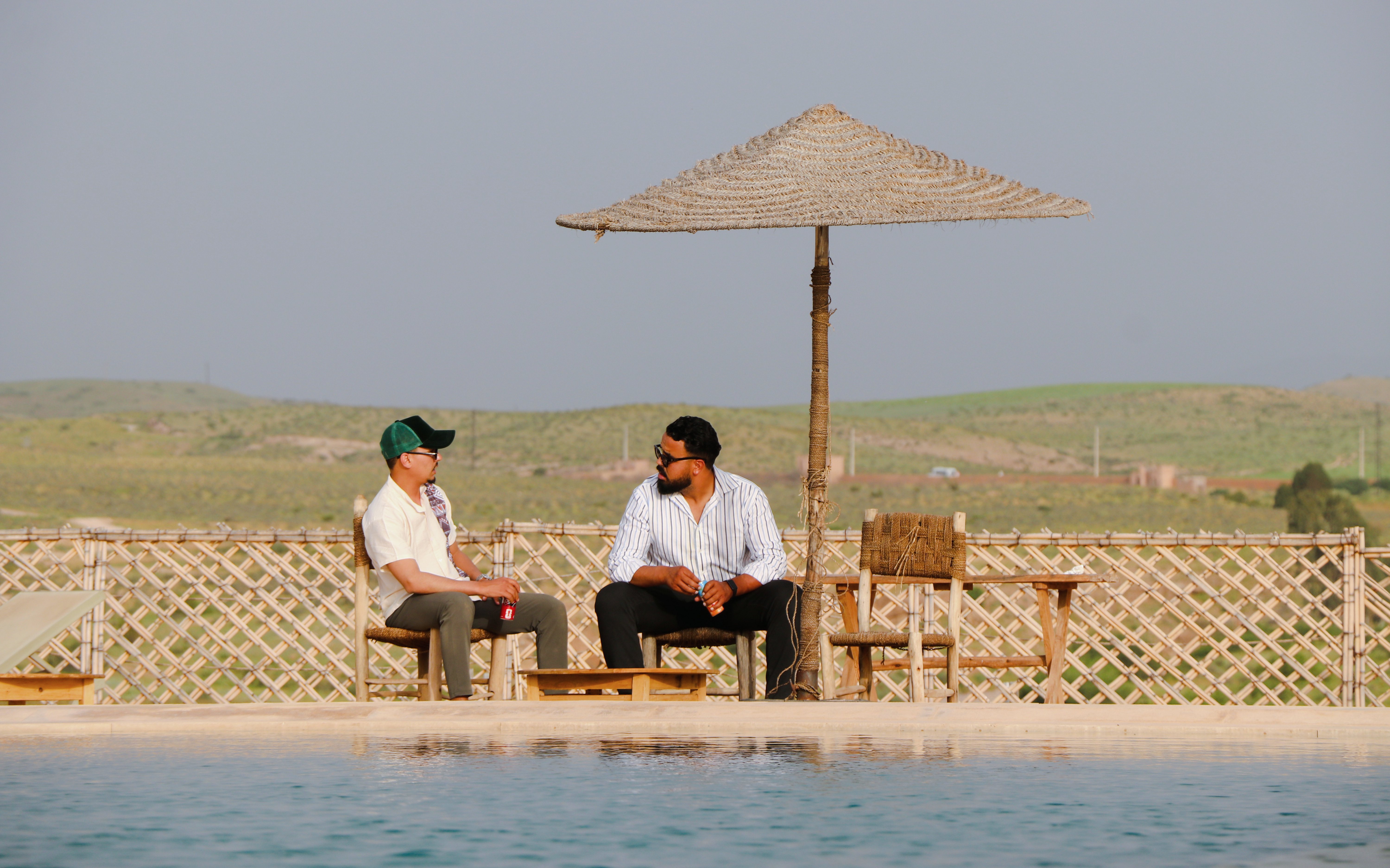 Two people sitting by a pool under a straw umbrella in Agafay desert.