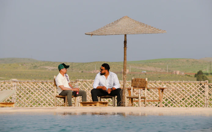 Two people sitting by a pool under a straw umbrella in Agafay desert.