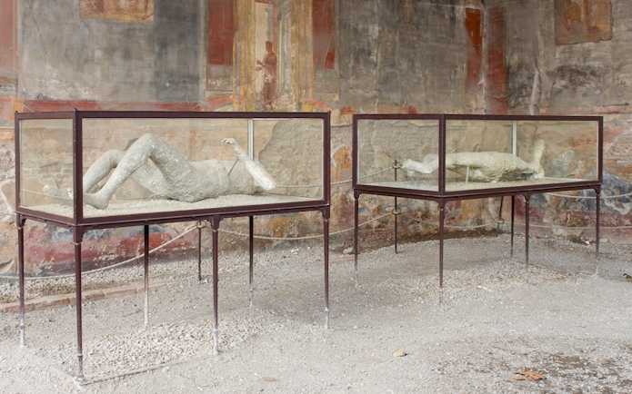 Victims of Pompeii eruption displayed in glass cases with ancient frescoes in the background.