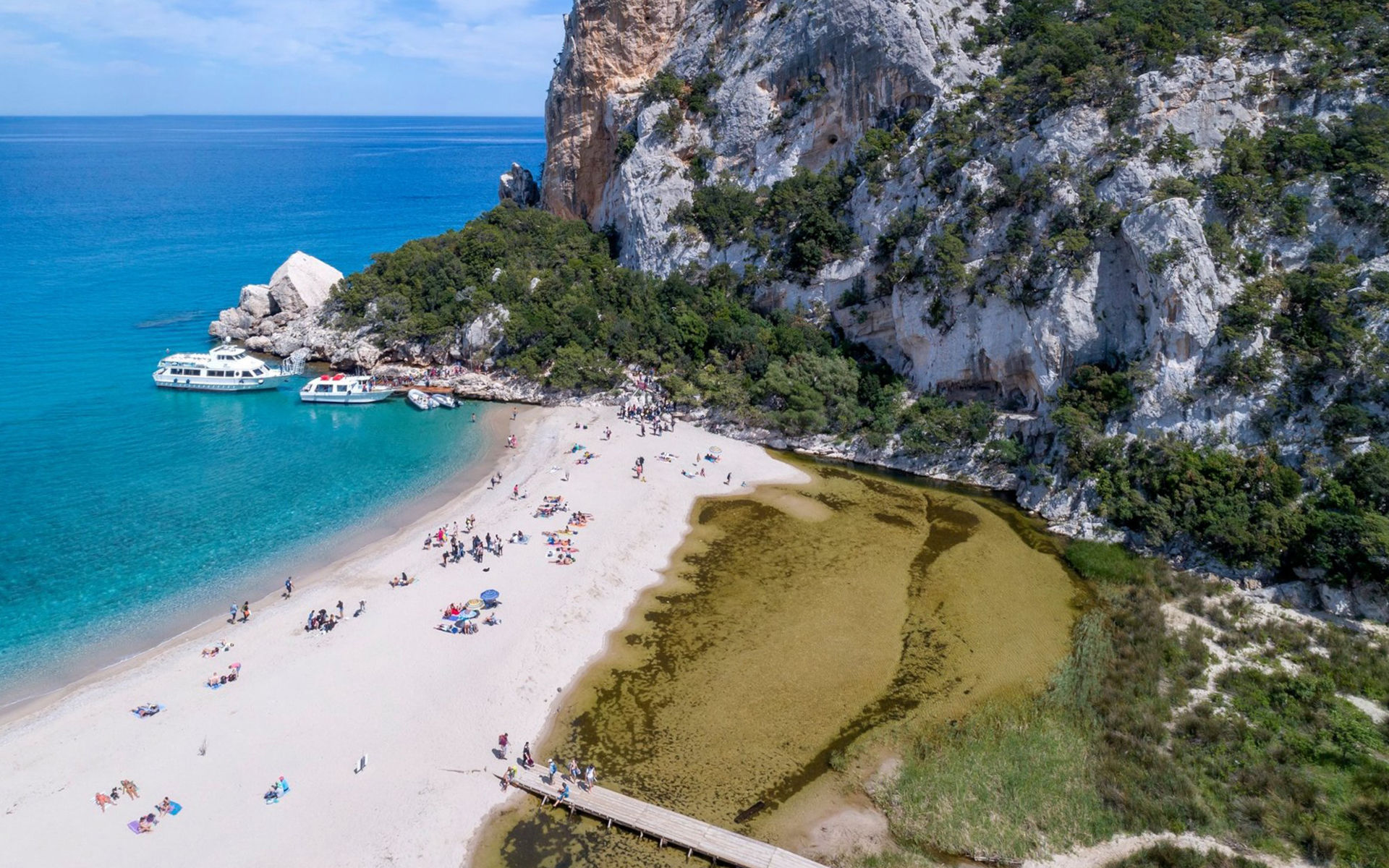 Beachgoers at Gulf of Orosei, Italy with boats and rocky cliffs in the background.