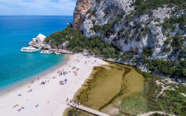 Beachgoers at Gulf of Orosei, Italy with boats and rocky cliffs in the background.