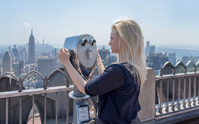Person using binoculars at Top of the Rock Observation Deck, New York City skyline in view.