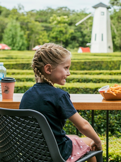 Children enjoying snacks at a table with a hedge maze and windmill in Amaze World.