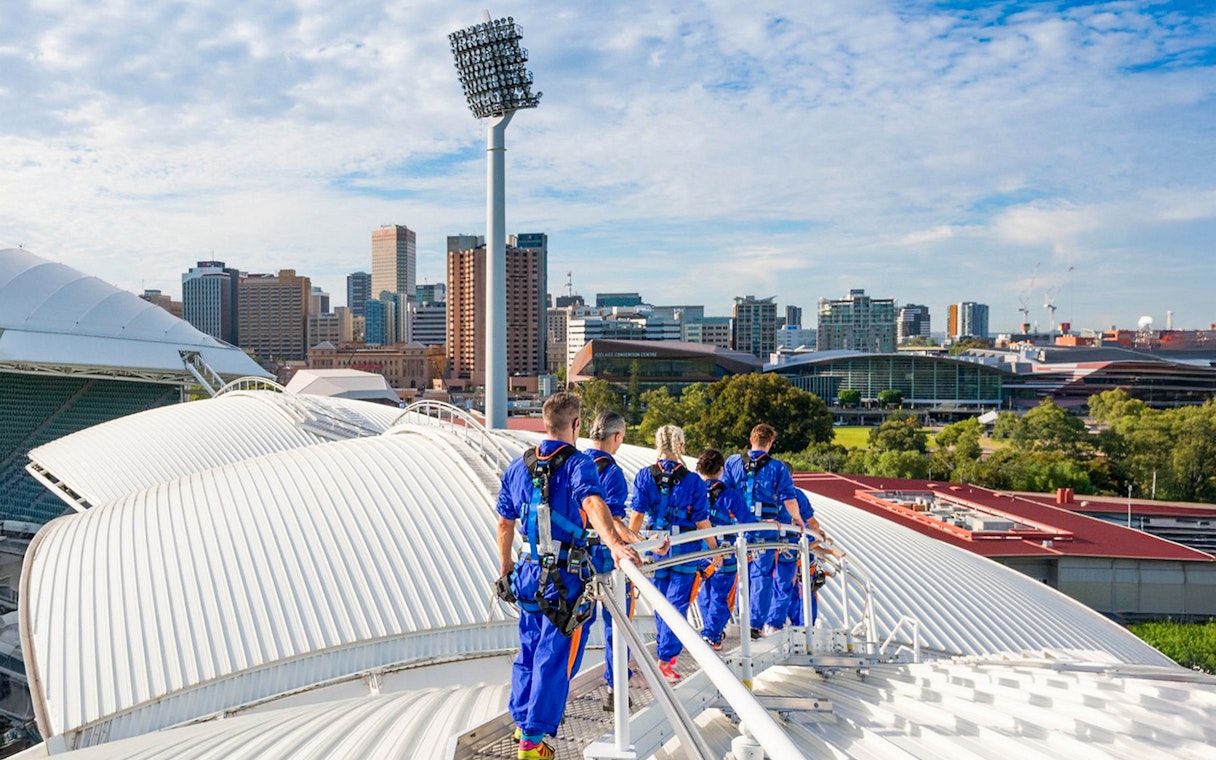 Group climbing Adelaide Oval rooftop with city skyline in background.