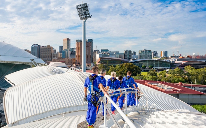 Group climbing Adelaide Oval rooftop with city skyline in background.