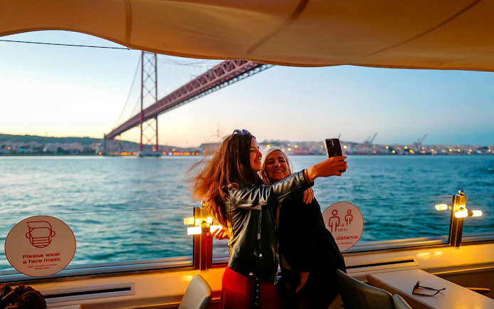 Two people taking a selfie on a sunset cruise on the Tagus River in Lisbon with a bridge in the background.