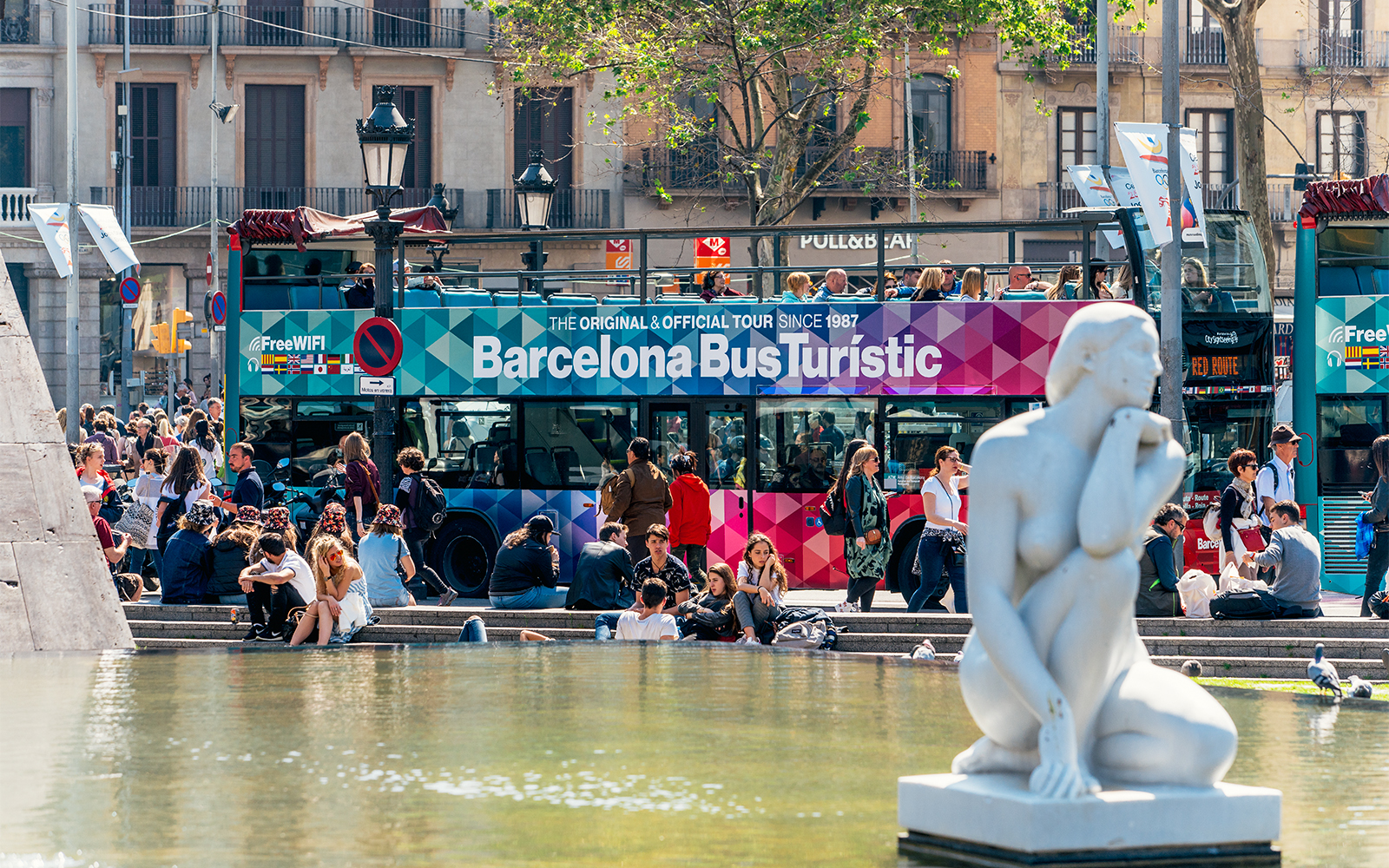 Barcelona Bus Turistic at a busy plaza with a white statue in the foreground.