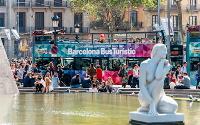 Barcelona Bus Turistic at a busy plaza with a white statue in the foreground.