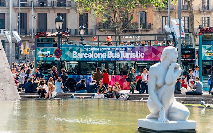 Barcelona Bus Turistic at a busy plaza with a white statue in the foreground.
