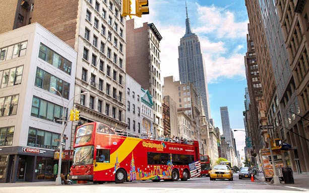 Open-top sightseeing bus on New York City street with Empire State Building in background.