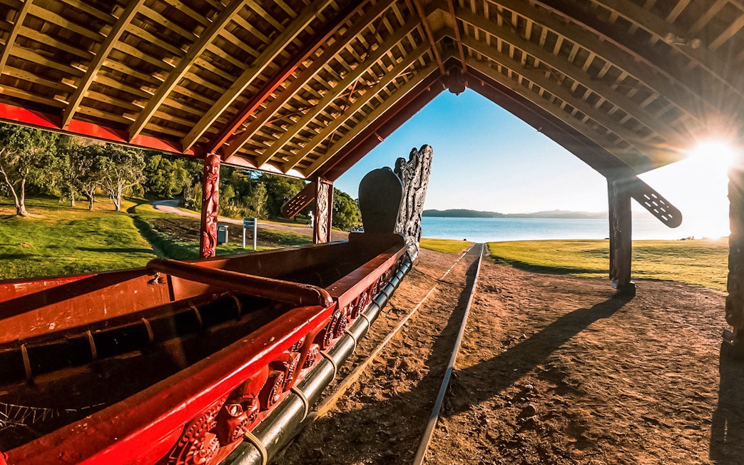 Ngtokimatawhaorua waka taua under shelter at sunrise by a lake.