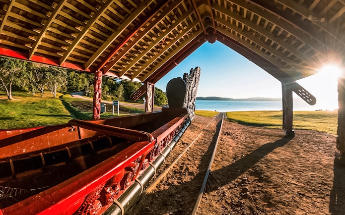 Ngtokimatawhaorua waka taua under shelter at sunrise by a lake.