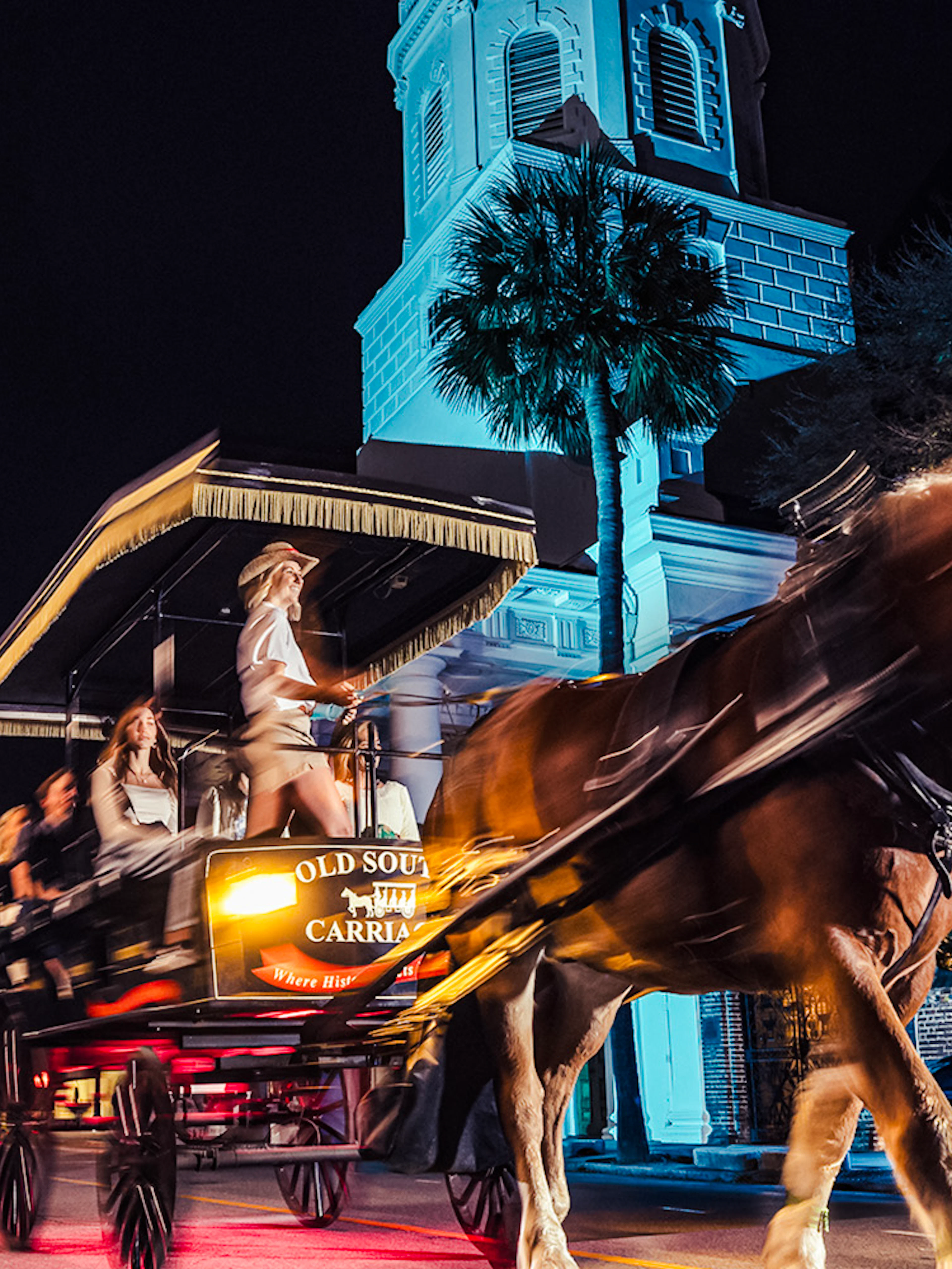 Horse-drawn carriage tour at night in historic Charleston, passing a lit church.