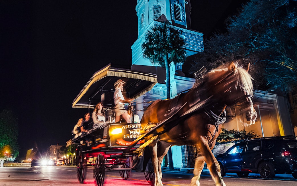 Horse-drawn carriage tour at night in historic Charleston, passing a lit church.