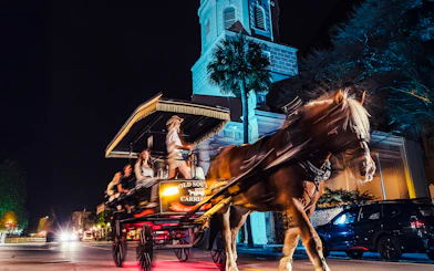 Horse-drawn carriage tour at night in historic Charleston, passing a lit church.
