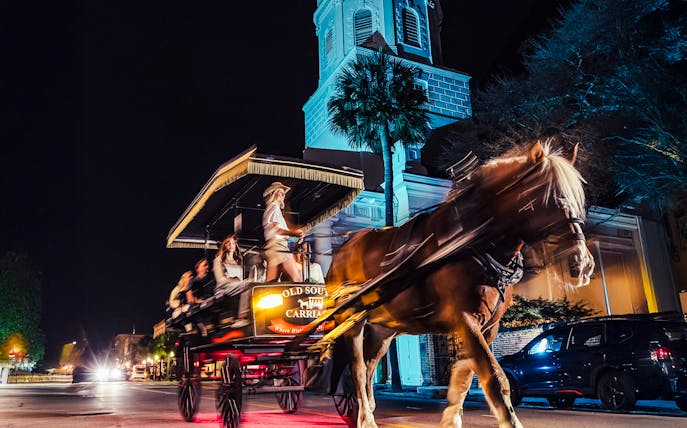 Horse-drawn carriage tour at night in historic Charleston, passing a lit church.