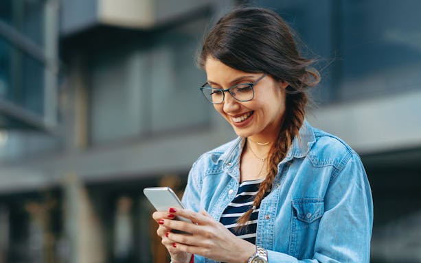Woman smiling while reading a message on her phone outdoors.