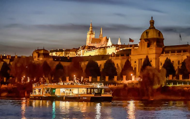 Vltava River cruise boat with Prague Castle illuminated at dusk.