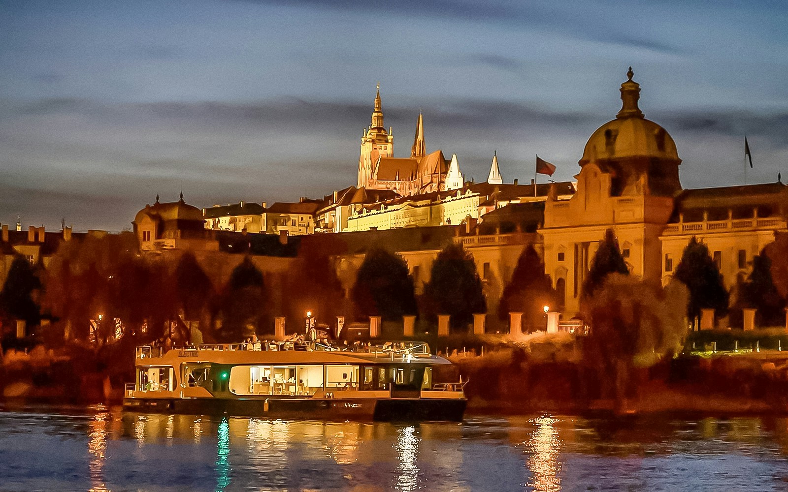 Vltava River cruise boat with Prague Castle illuminated at dusk.