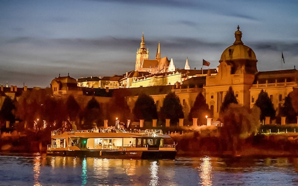 Vltava River cruise boat with Prague Castle illuminated at dusk.