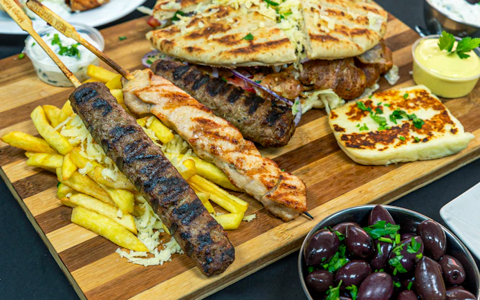 Grilled meats and pita bread with fries, olives, and halloumi on a wooden platter, Greek cuisine.