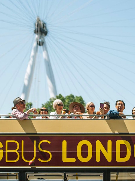 Open-top Big Bus tour in London with tourists, London Eye in background.