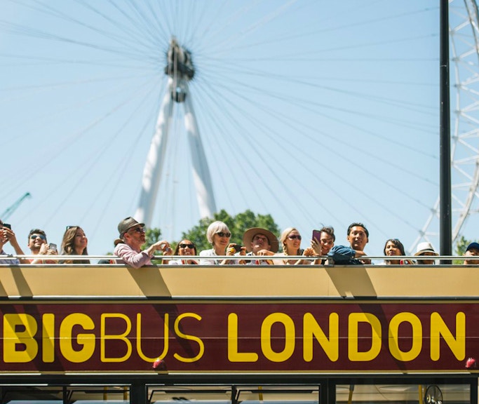 Open-top Big Bus tour in London with tourists, London Eye in background.