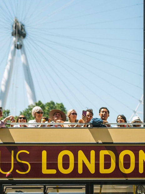 Open-top Big Bus tour in London with tourists, London Eye in background.