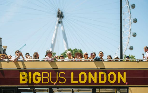 Open-top Big Bus tour in London with tourists, London Eye in background.