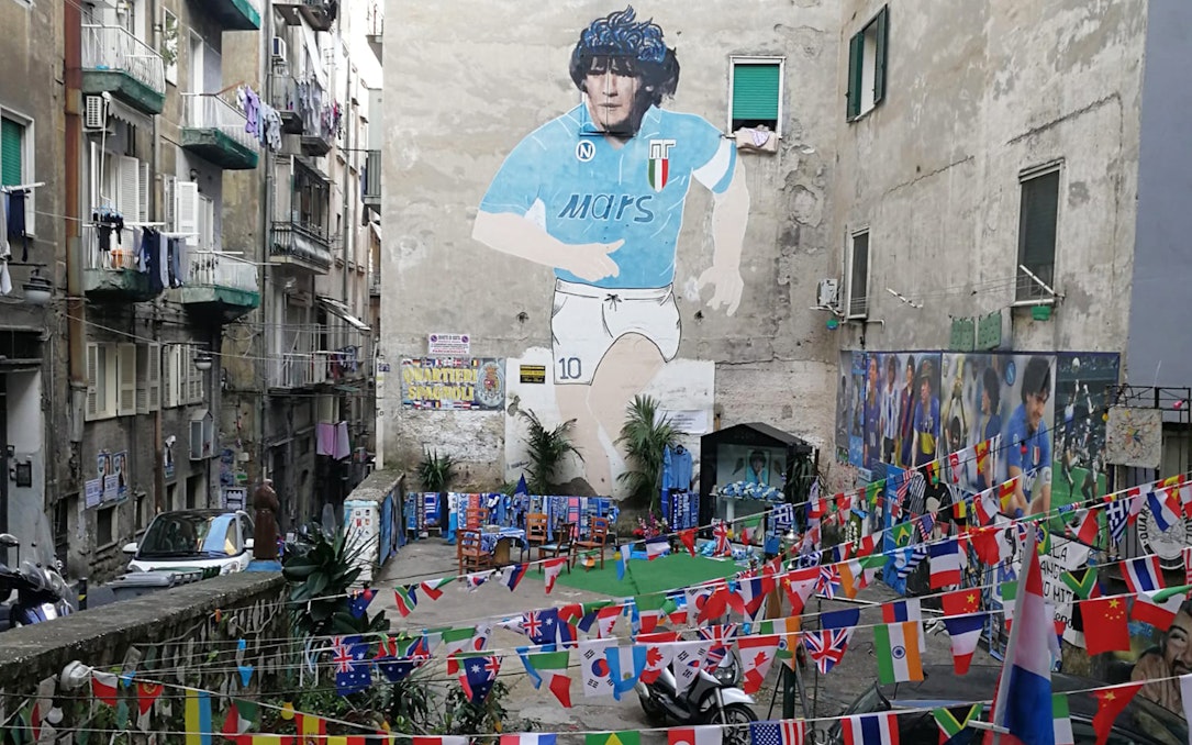 Mural of Maradona in Naples alley with international flags and street art.