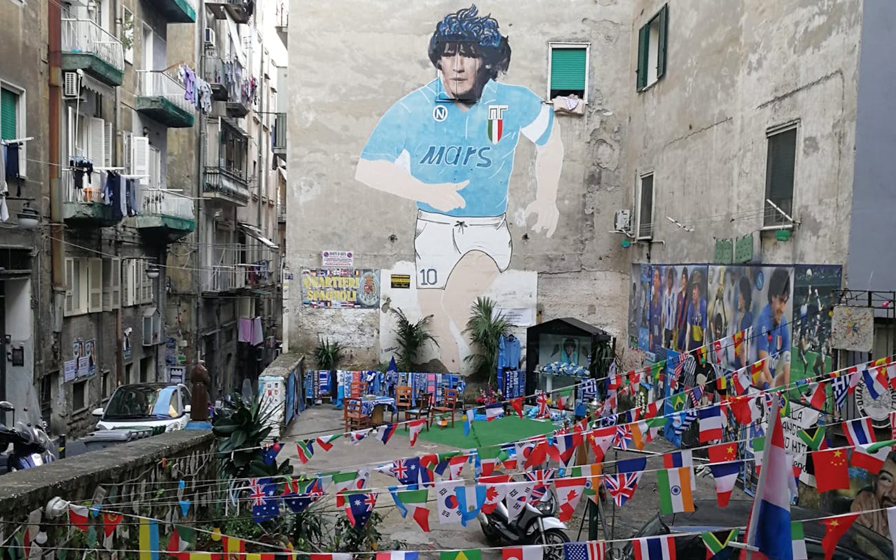 Mural of Maradona in Naples alley with international flags and street art.