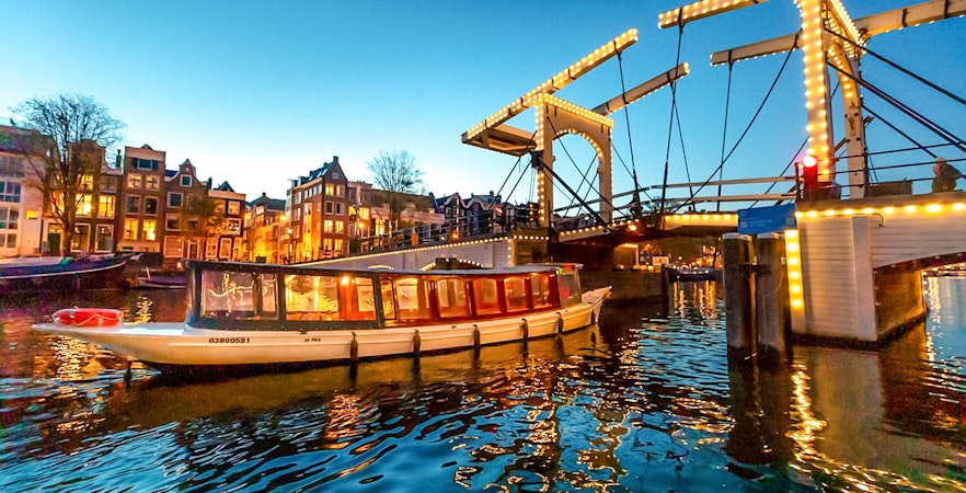 Canal boat cruising under illuminated bridge during Amsterdam Light Festival.