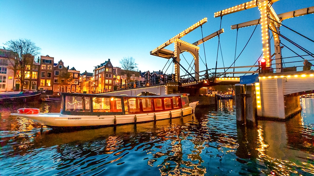 Canal boat cruising under illuminated bridge during Amsterdam Light Festival.