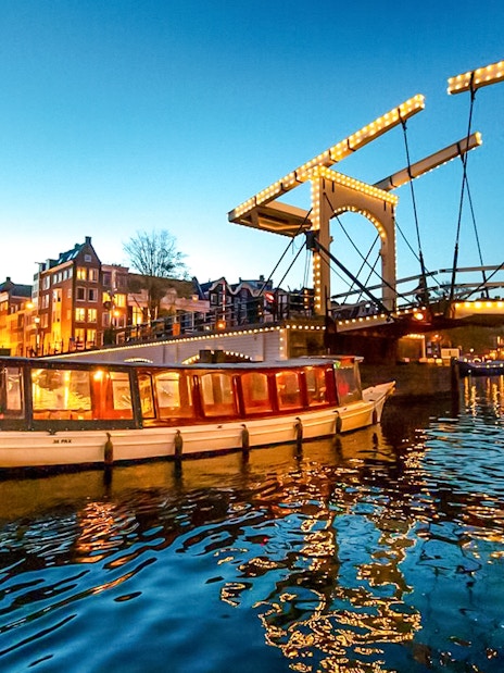Canal boat cruising under illuminated bridge during Amsterdam Light Festival.
