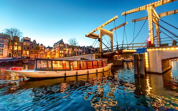 Canal boat cruising under illuminated bridge during Amsterdam Light Festival.