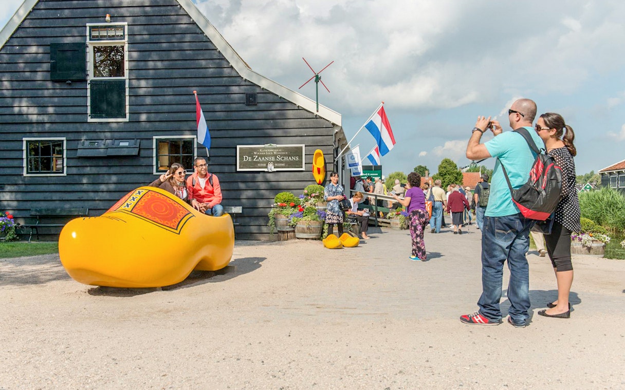 Visitors at Zaanse Schans posing in a giant wooden shoe with Dutch flags and windmill in the background.