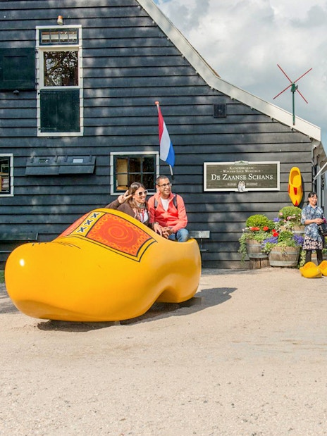 Visitors at Zaanse Schans posing in a giant wooden shoe with Dutch flags and windmill in the background.