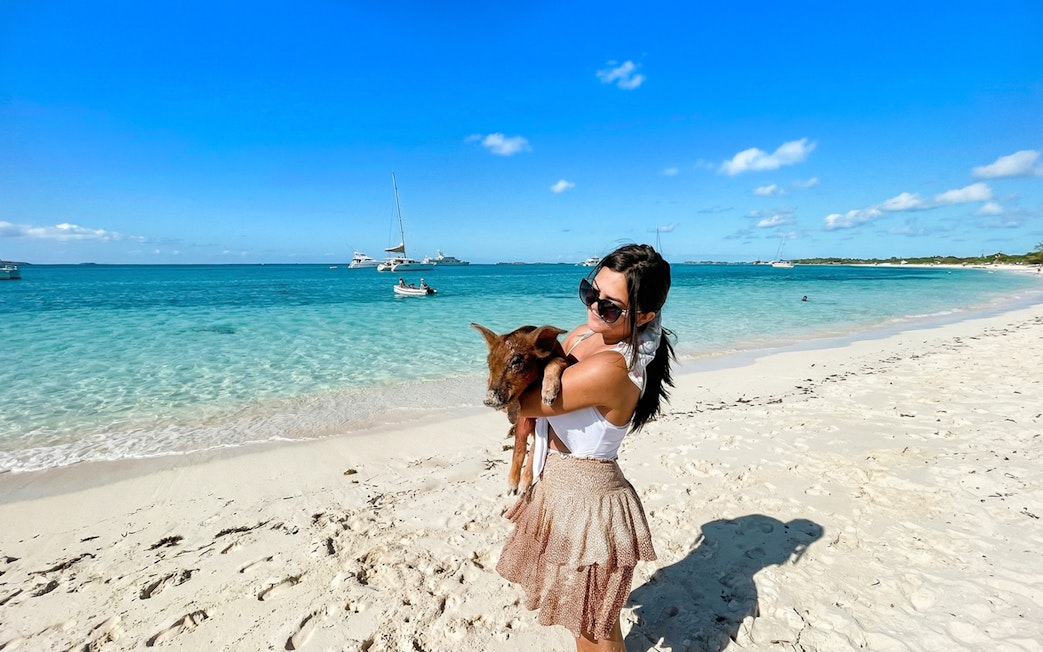 Woman holding a pig on a sandy beach in Exuma, Bahamas with boats in the background.