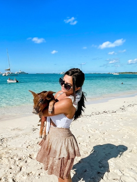 Woman holding a pig on a sandy beach in Exuma, Bahamas with boats in the background.