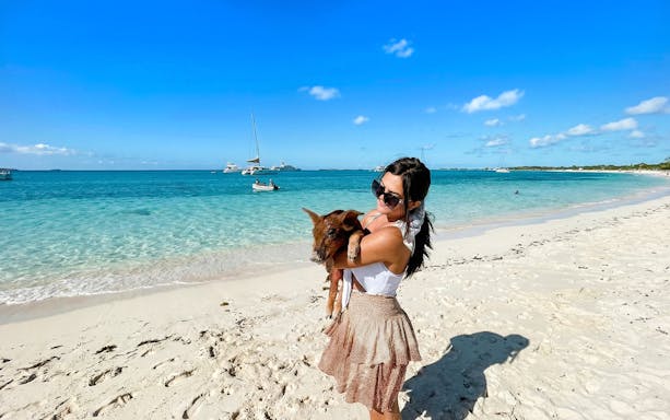 Woman holding a pig on a sandy beach in Exuma, Bahamas with boats in the background.