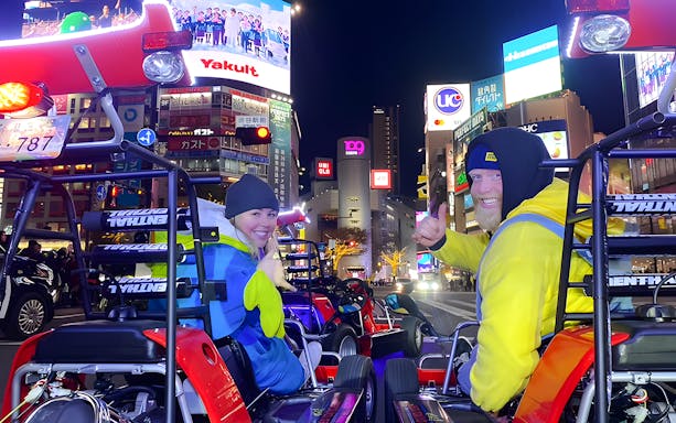 People in go-karts posing in Shibuya at night with bright city lights and billboards.