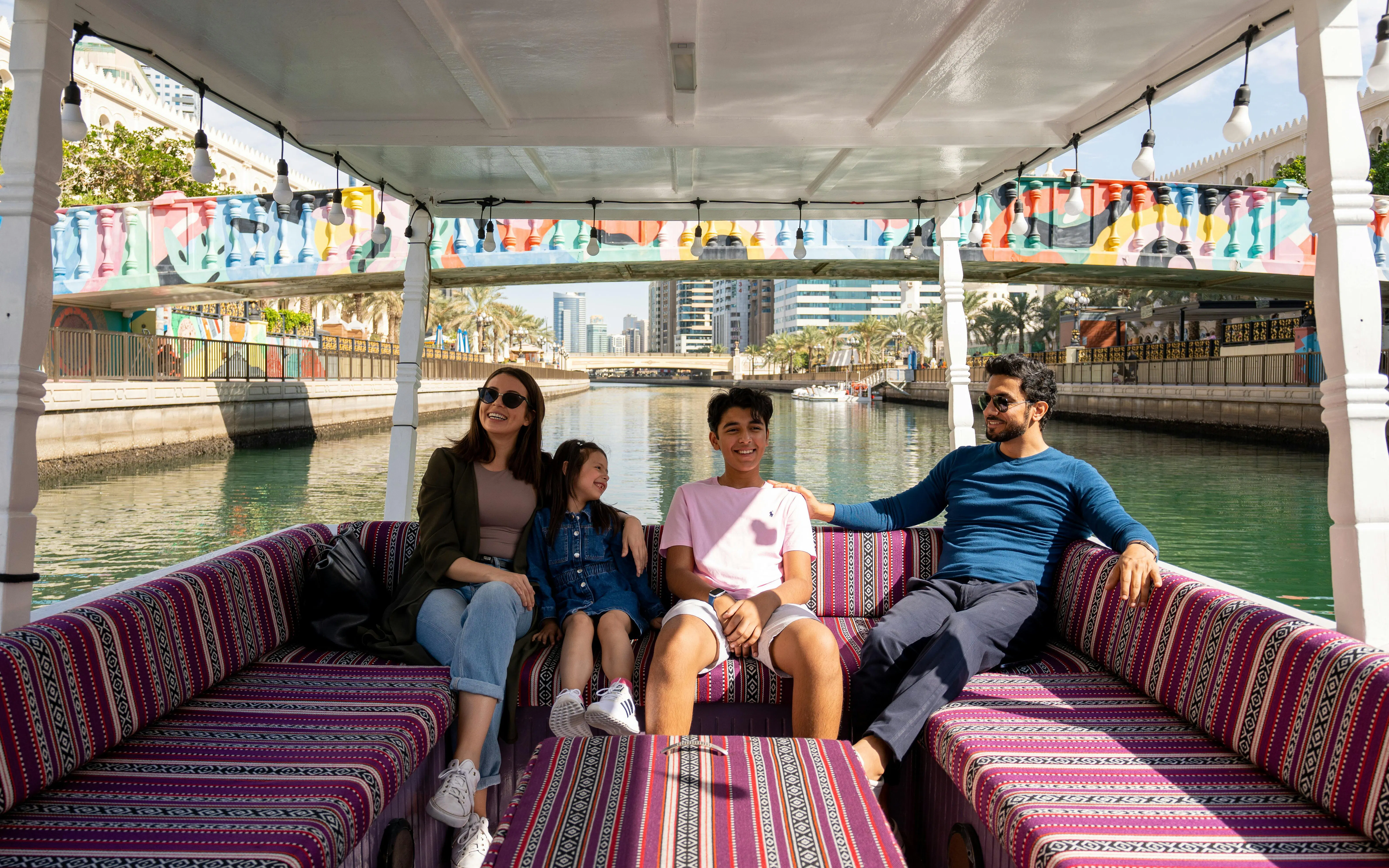Family enjoying a boat tour on a canal in Sharjah with colorful bridge in background.