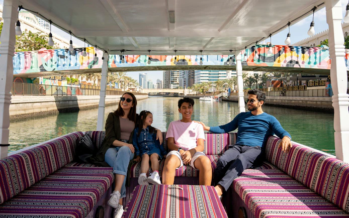 Family enjoying a boat tour on a canal in Sharjah with colorful bridge in background.