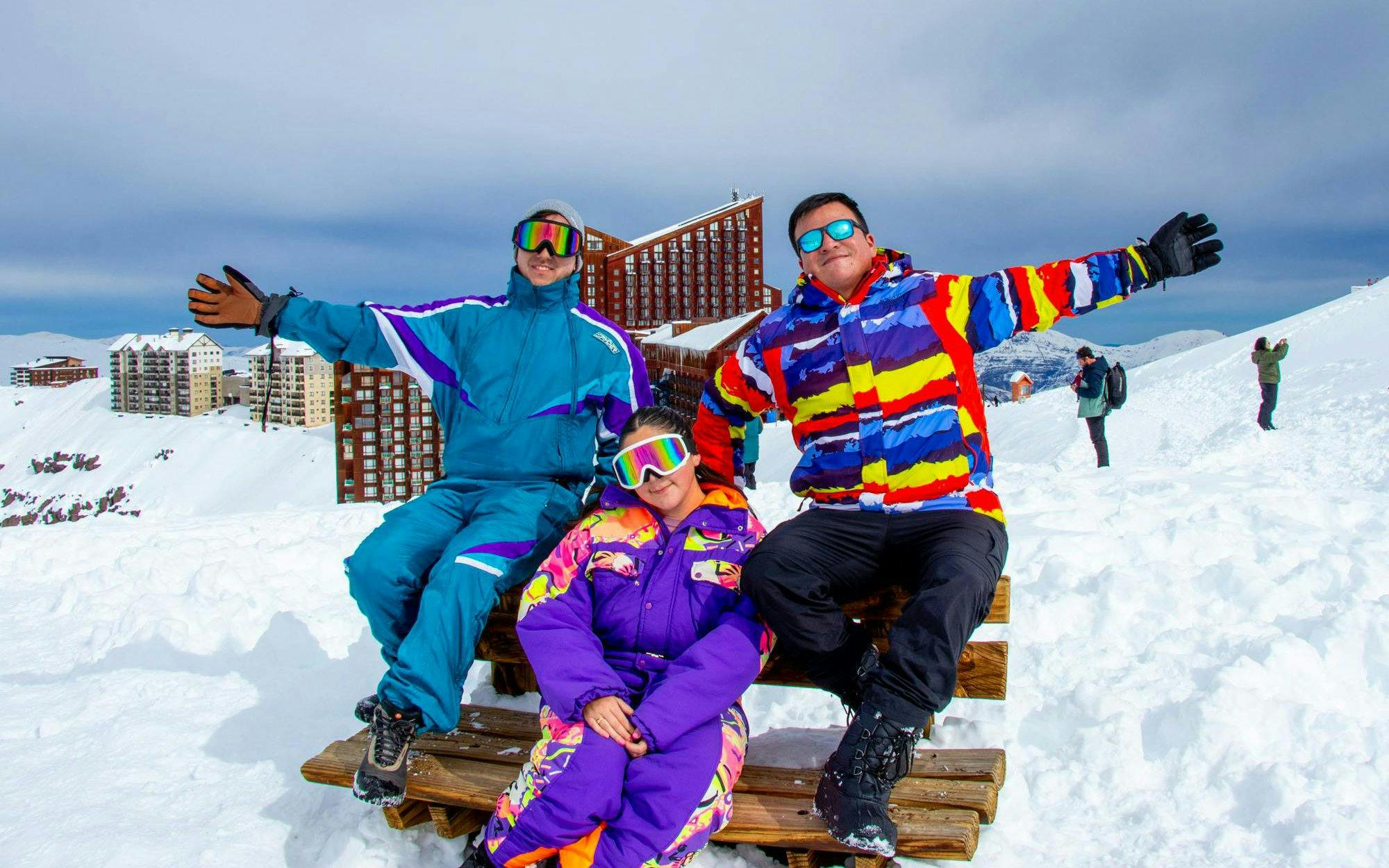 Tourists in colorful ski gear at Valle Nevado with snowy mountains and buildings in the background.