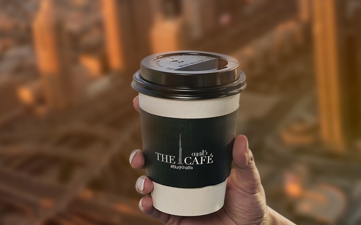 Hand holding a coffee cup from The Café at Burj Khalifa with city view in background.